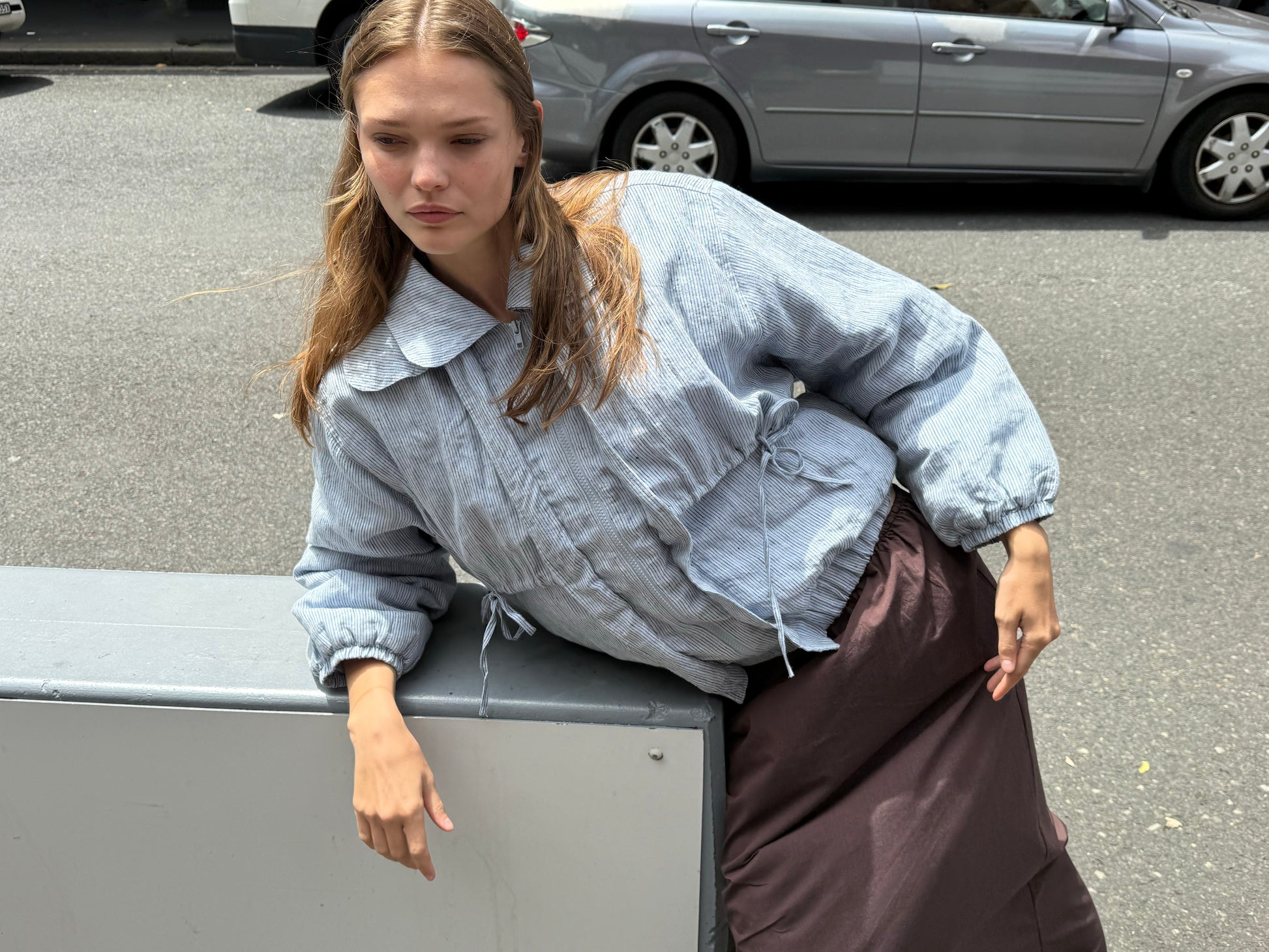 A woman with long, light brown hair leans against a metal barrier outdoors, wearing the Double Layered Jacket - Blue Stripe and a long brown skirt. Parked cars and a paved street are visible in the background.