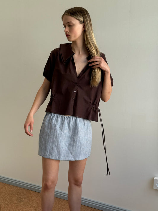 A woman stands indoors against a plain wall, wearing a loose dark brown short-sleeve shirt with a large collar and the Babylock Skirt - Blue Stripe. She looks to the side and touches her hair with one hand.