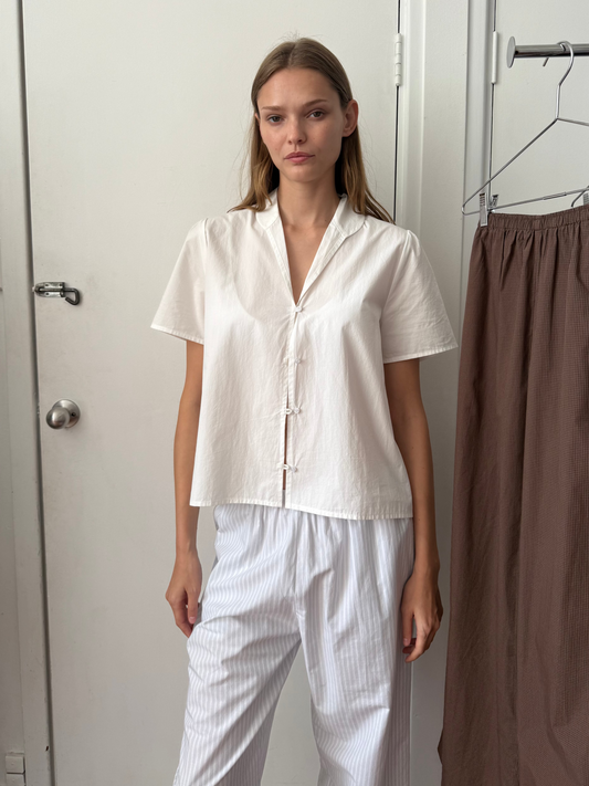 A woman stands indoors wearing the Sails Top - White with white striped pants. A brown skirt hangs on a nearby rack, and a white door and wall are in the background.