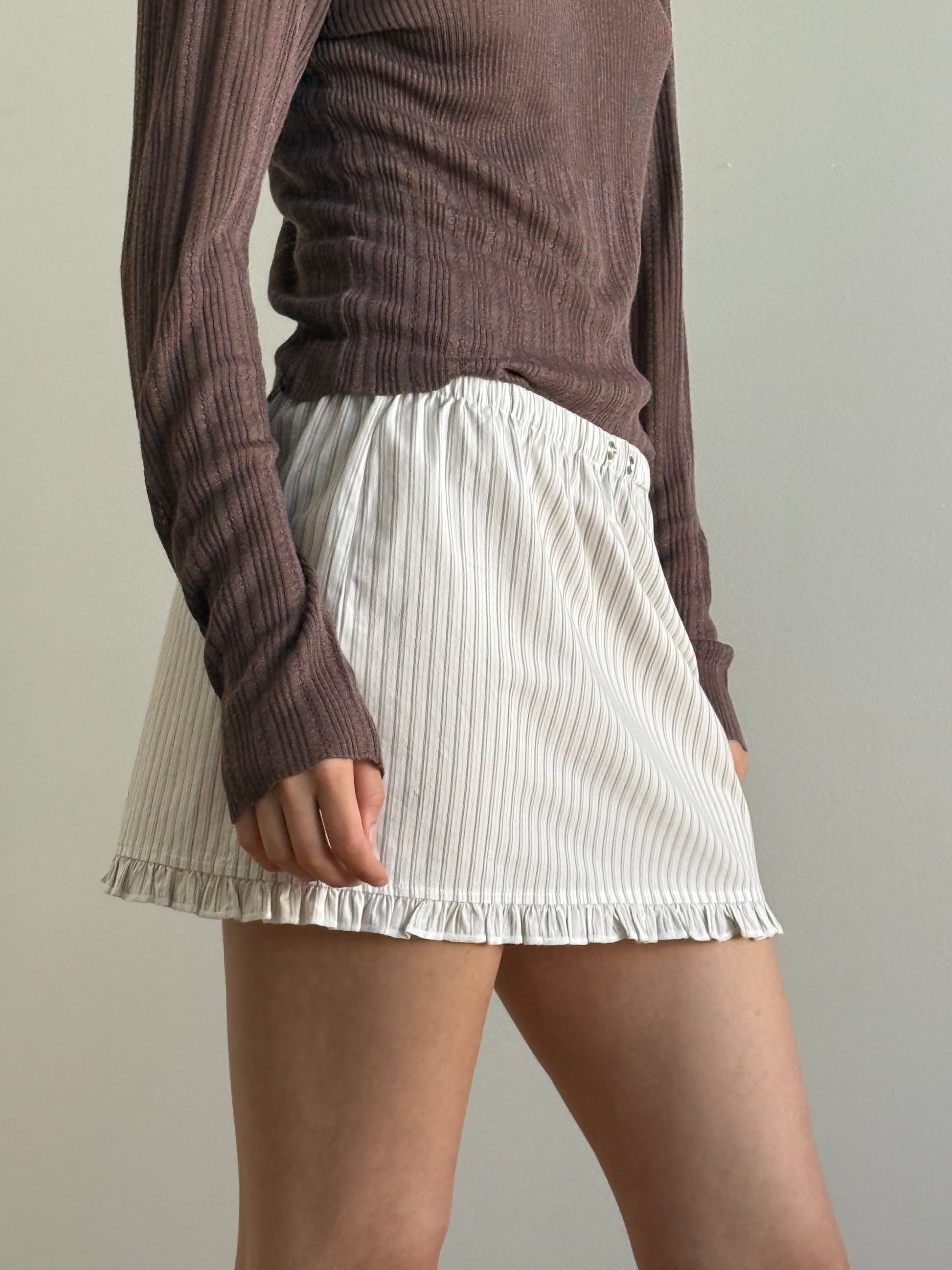 Someone stands against a plain light background, wearing a long-sleeved, ribbed brown top and the Flounce Mini - Gull Stripe by Deiji Studios—an organic cotton skirt with subtle vertical stripes and a ruffled hem.