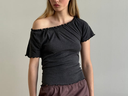 A woman wears the Flounce Tee - Grey by Deiji Studios with a mauve skirt, standing against a plain light grey background. Her hair is down and her face is partially out of frame.