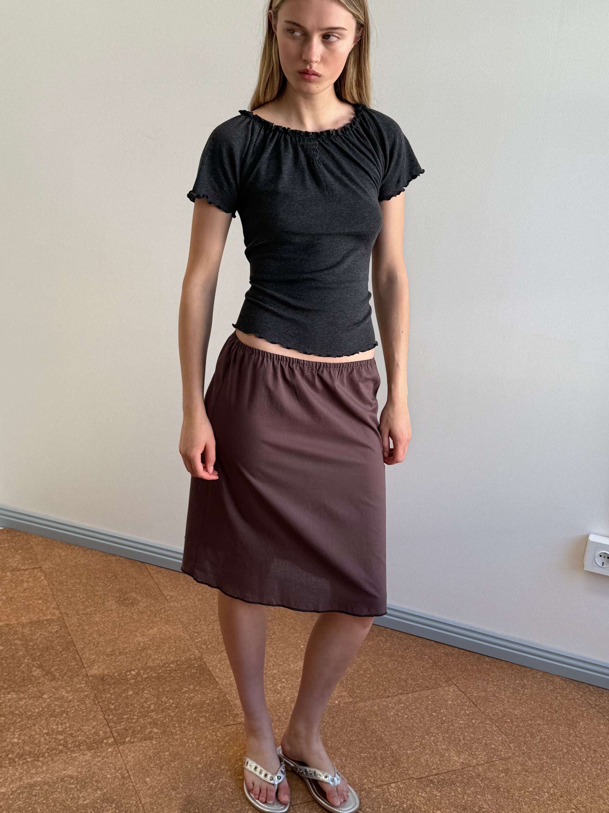 A woman stands indoors wearing a fitted black top, sandals, and the Simple Skirt - Raisin. Her loose brown organic cotton skirt falls to her knees as she looks slightly to the side against a plain light-colored wall.