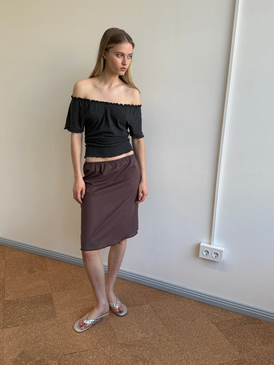 A woman with long blonde hair stands indoors against a plain wall, wearing an off-the-shoulder black top, the Simple Skirt in Raisin, and silver sandals. She looks slightly to the side with a neutral expression.