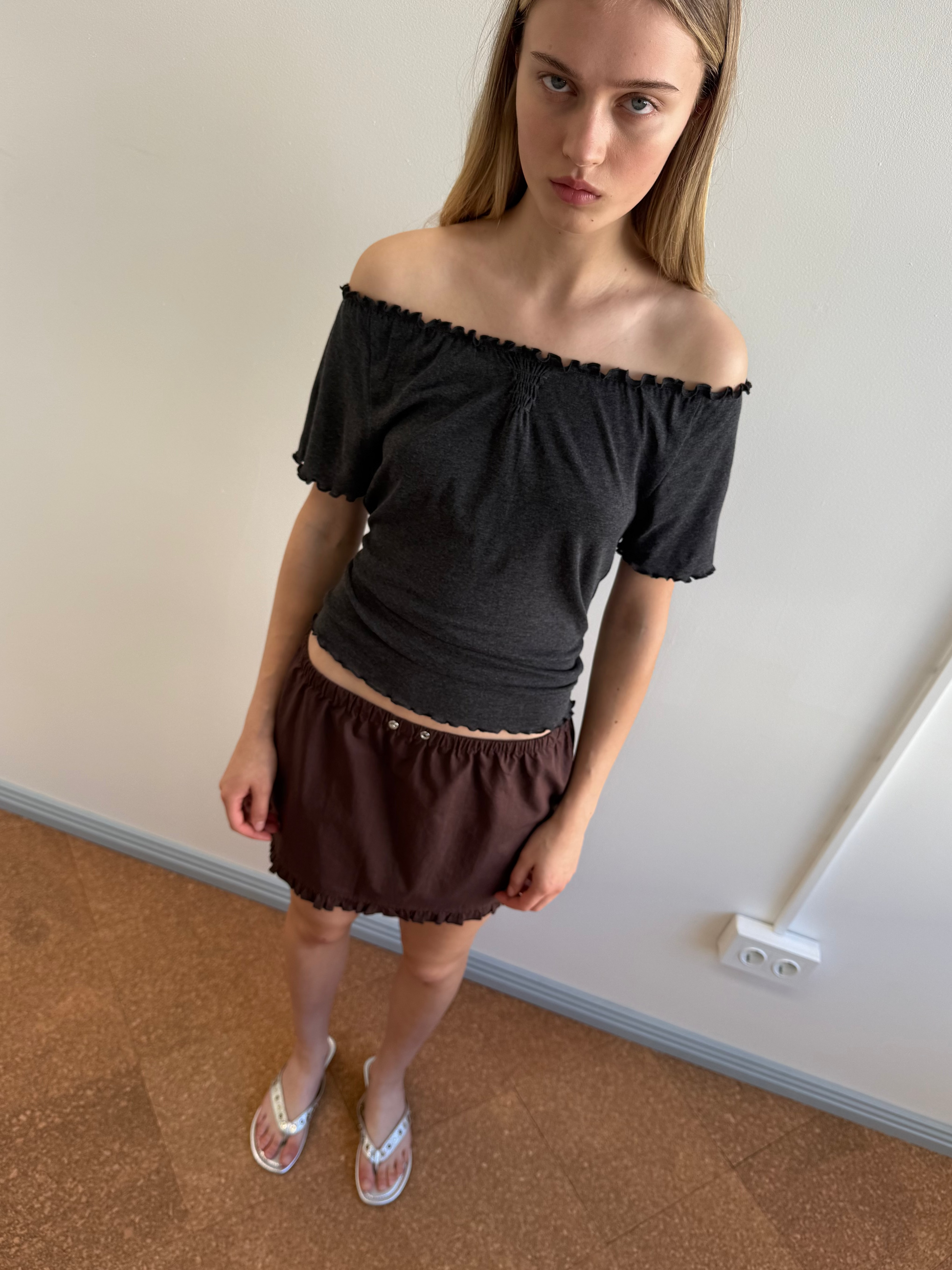 A young woman with long blonde hair stands indoors against a plain wall, wearing the Deiji Studios Flounce Mini - Umber paired with a dark off-shoulder top and white flip-flops, gazing upward with a neutral expression.