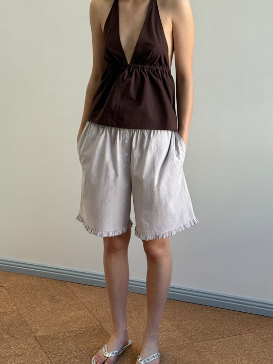 A person stands indoors wearing a sleeveless dark brown halter top and Deiji Studios' Flounce Short in Lilac Stripe, hands in pockets with white sandals, posing against a plain background with a blue baseboard.
