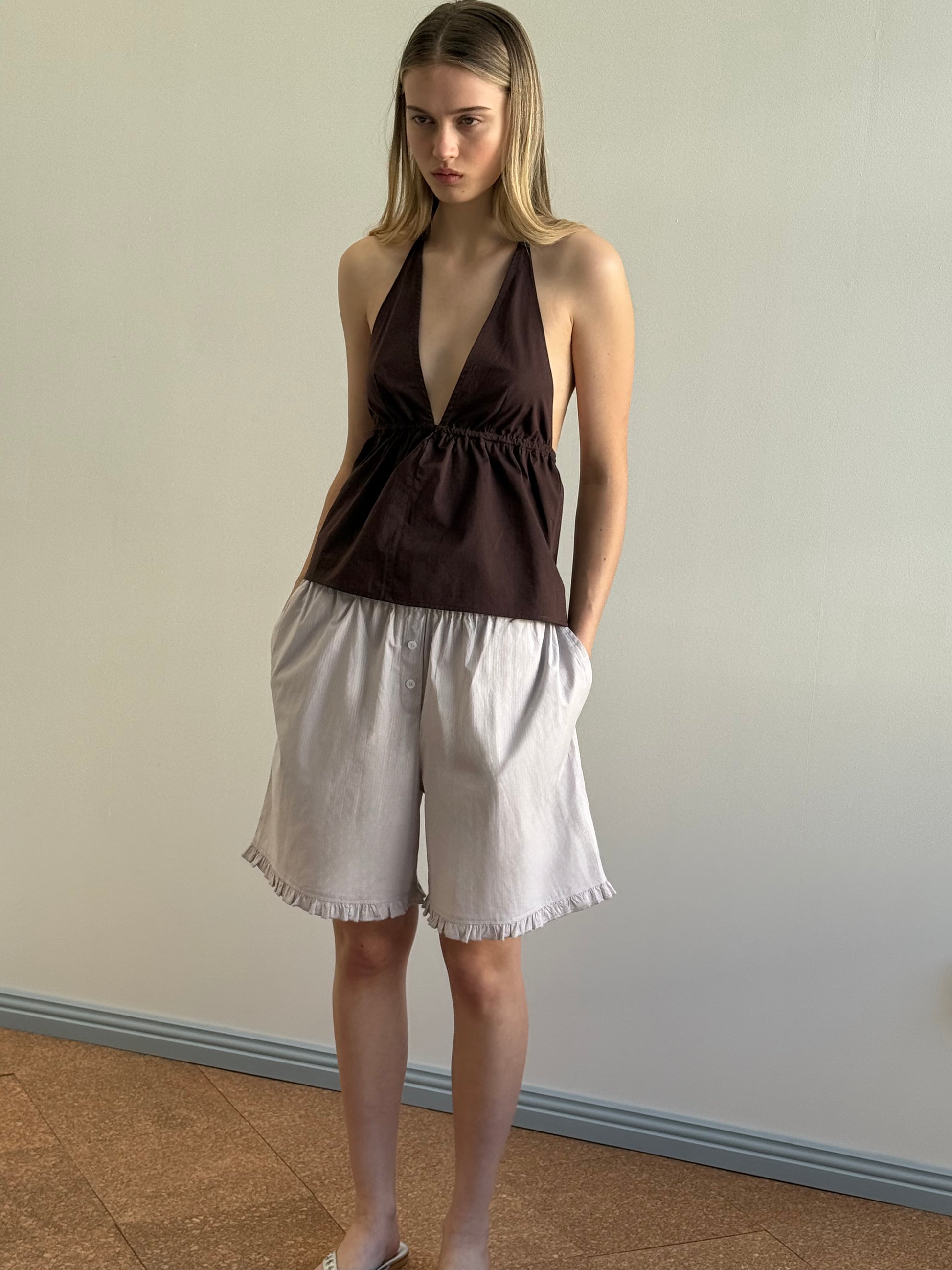 A woman with straight light brown hair and a neutral expression stands indoors against a plain wall, wearing a sleeveless dark brown halter top and relaxed fit Flounce Short - Lilac Stripe featuring ruffled hems.