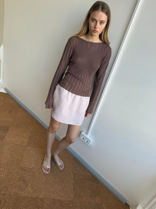 A young woman with straight blonde hair stands indoors against a pale wall, wearing a long-sleeved brown ribbed top, silver sandals, and the Babylock Skirt in lilac. She looks slightly down with a neutral expression.