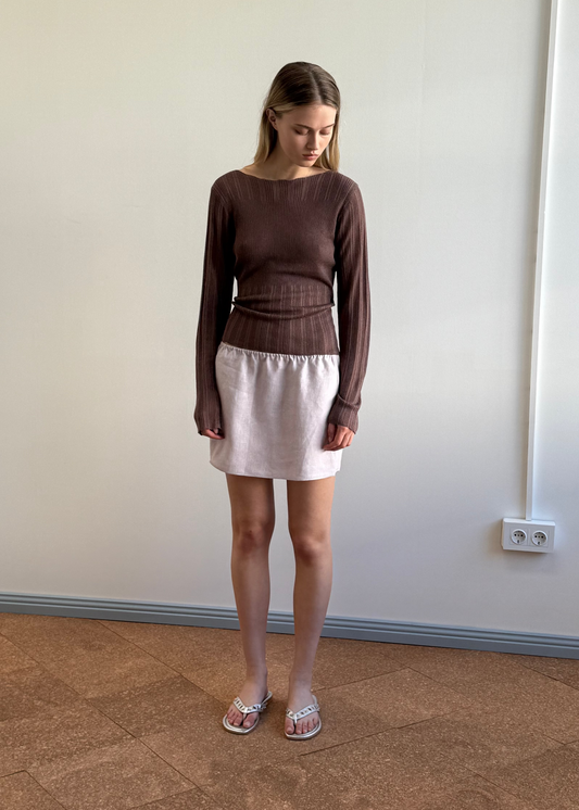 A young woman stands indoors in a long-sleeve brown top and the Babylock Skirt - Lilac, paired with white sandals, looking down with her hands by her sides against a plain wall with a visible power outlet.