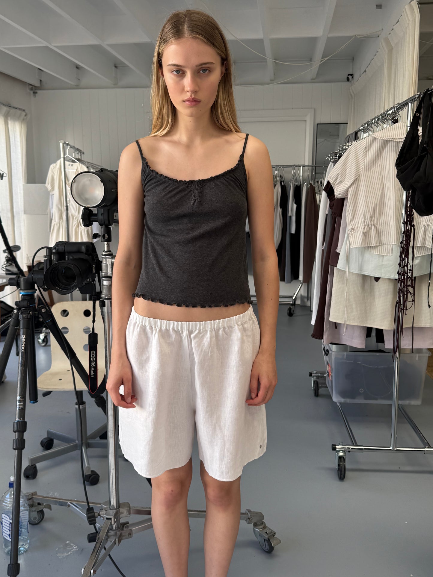A young woman with straight blonde hair stands indoors in a dark sleeveless top and Deiji Studios' Press Stud Short in White, surrounded by clothing racks and photography equipment in a well-lit studio.