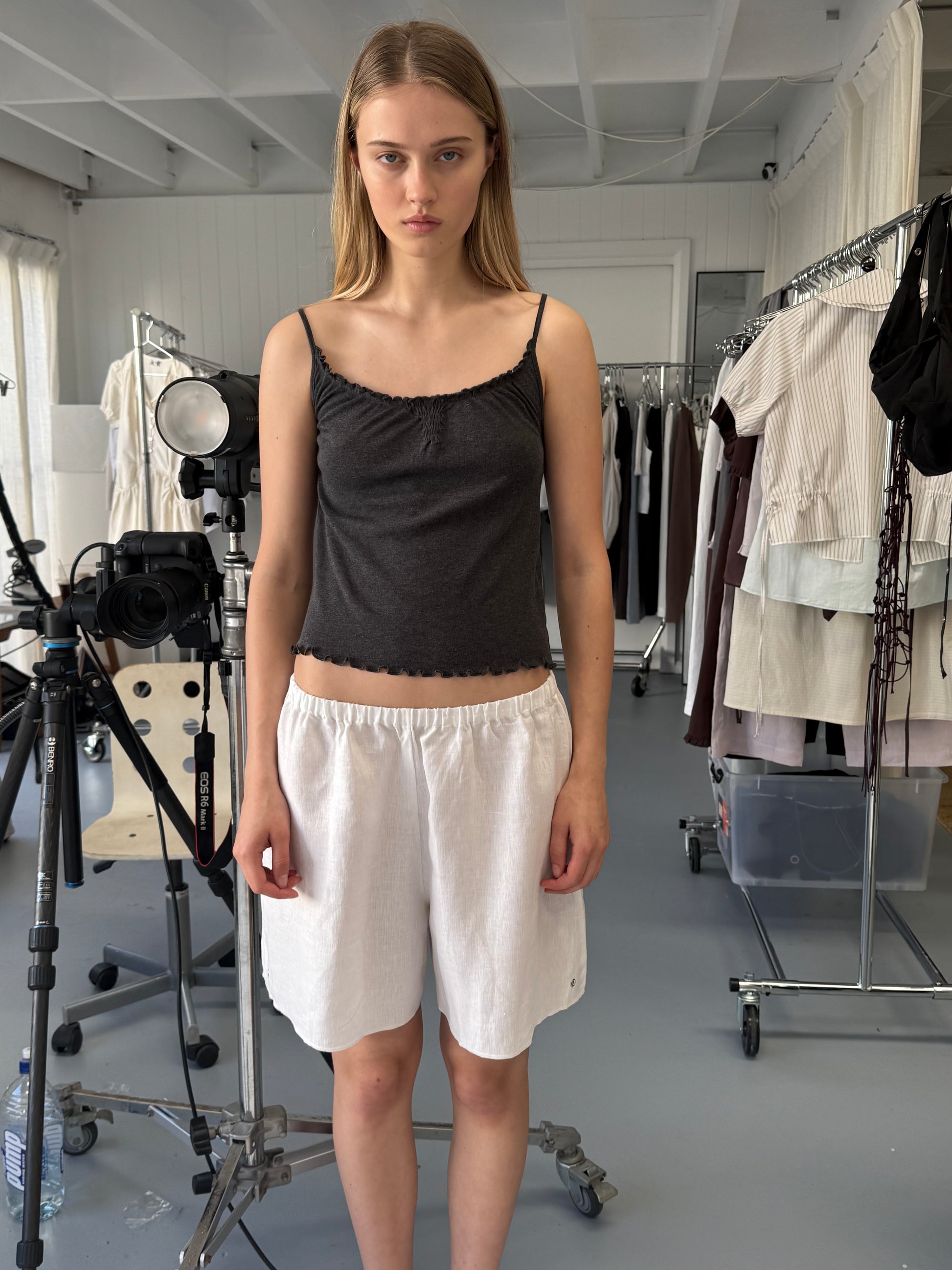 A young woman with straight blonde hair stands indoors in a dark sleeveless top and Deiji Studios' Press Stud Short in White, surrounded by clothing racks and photography equipment in a well-lit studio.