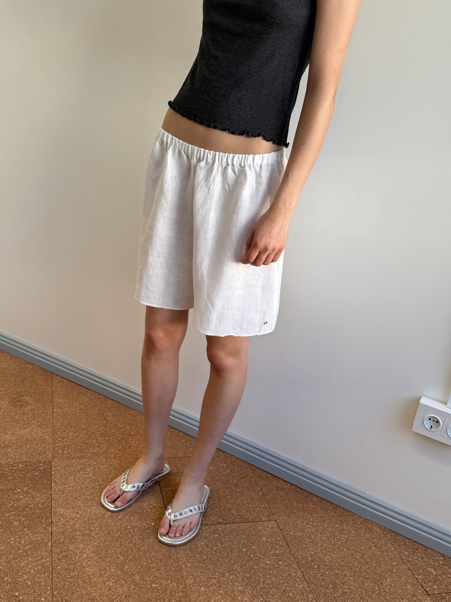 A person stands indoors on a brown floor next to a light wall with an outlet, wearing a black sleeveless top, Press Stud Short - White by Deiji Studios, and white flip-flops.