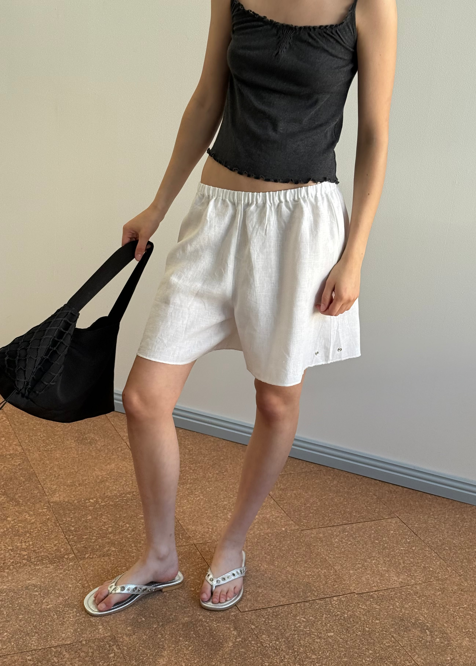 A person wears a black sleeveless top, Deiji Studios Press Stud Short - White, and white sandals, holding a black woven bag. They stand indoors on brown tiles with a plain light-colored wall behind them.