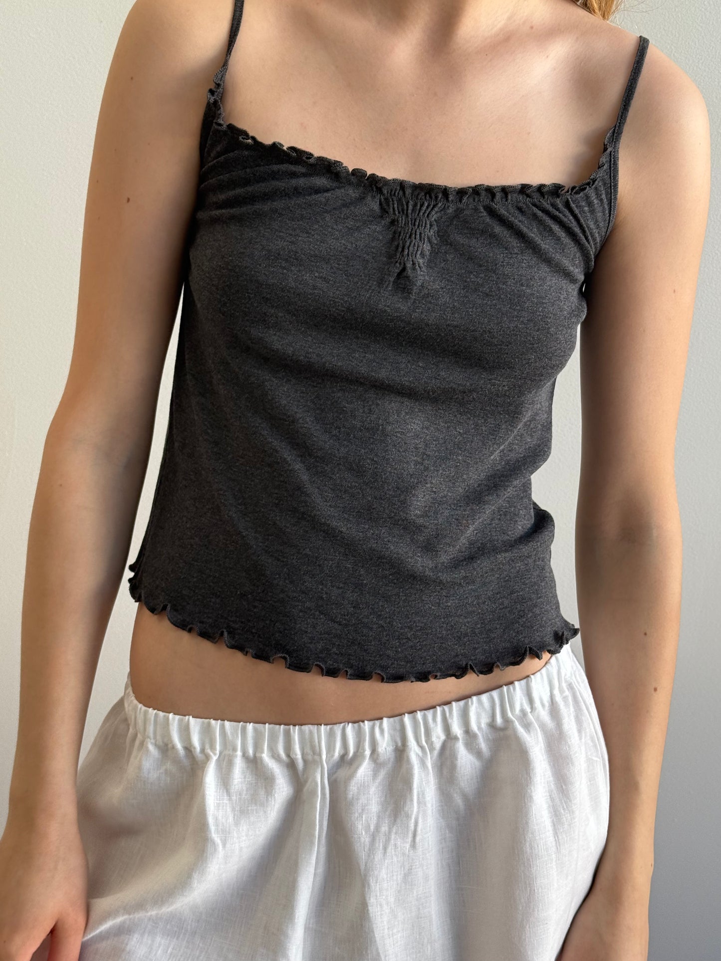 Wearing the Flounce Tank - Grey from Deiji Studios, a person pairs this dark grey, sleeveless top with ruffled edges and loose white shorts while standing against a plain light background.
