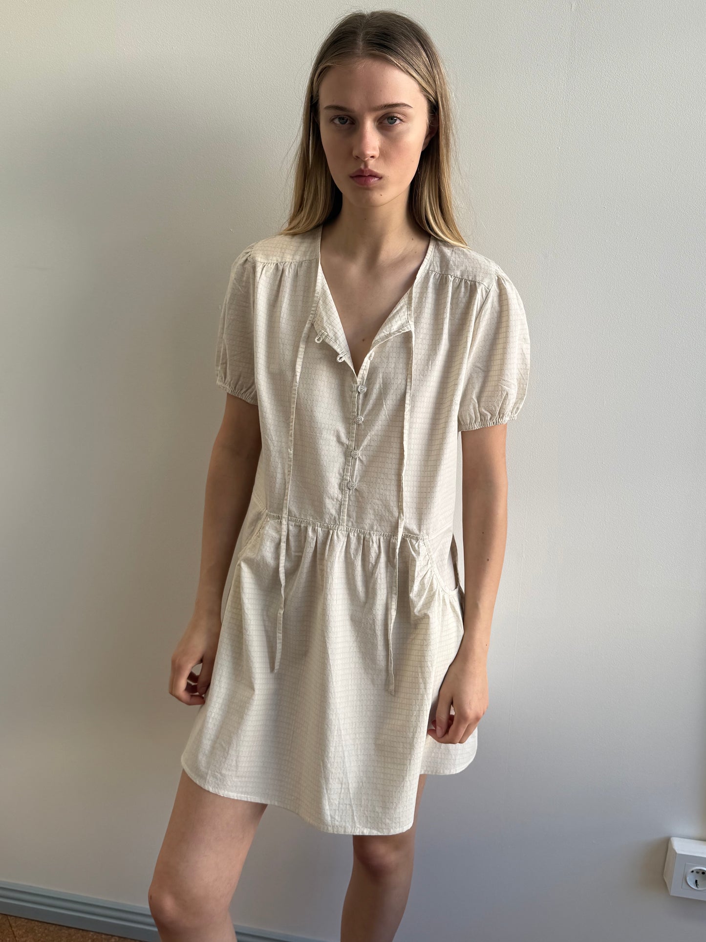 A young woman with long blonde hair stands indoors by a plain light wall, wearing the Deiji Studios Pocket Mini Dress in Bone Check—a short-sleeved, knee-length organic cotton style with a gathered waist and tie neckline.