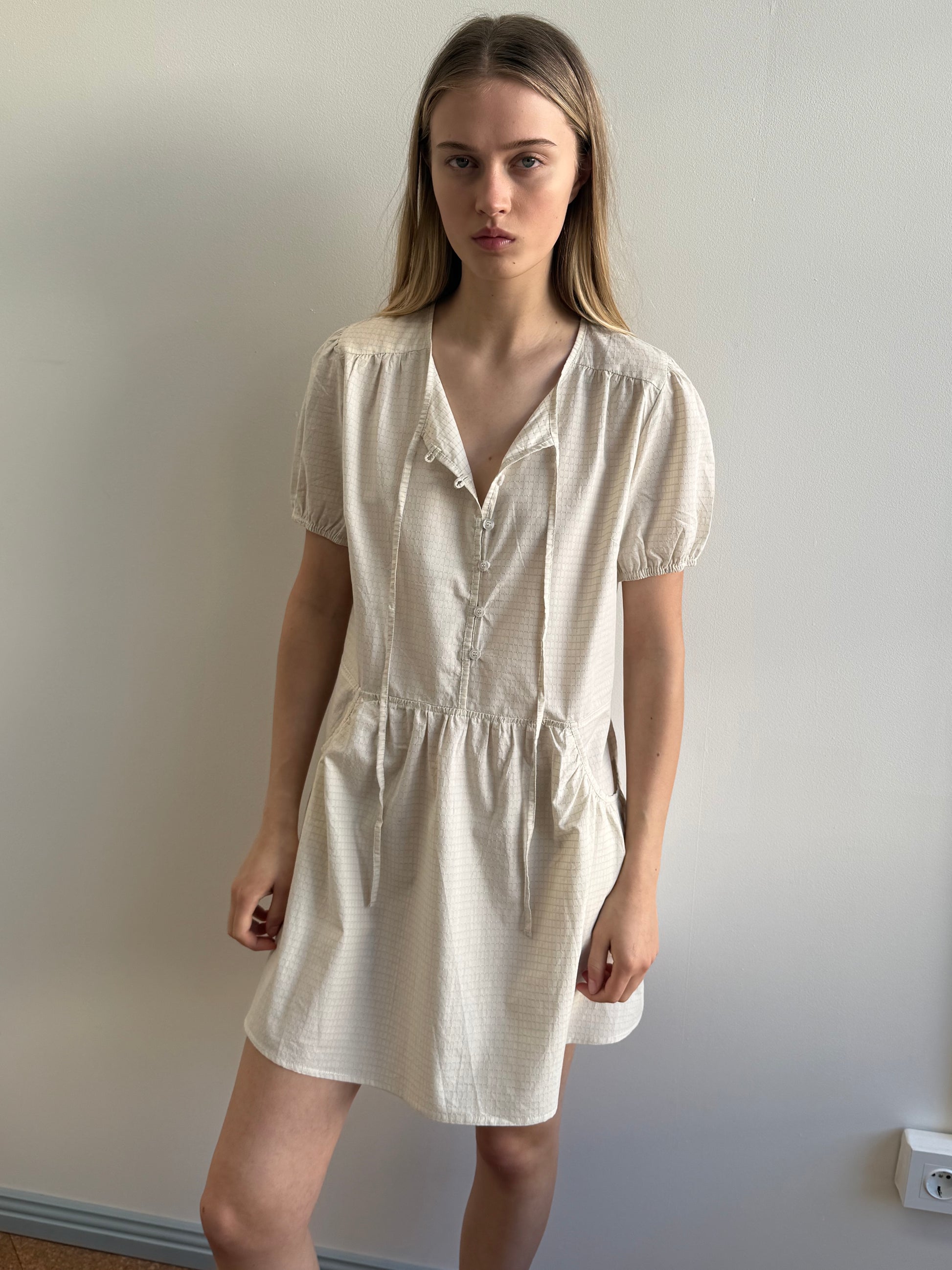 A young woman with long blonde hair stands indoors by a plain light wall, wearing the Deiji Studios Pocket Mini Dress in Bone Check—a short-sleeved, knee-length organic cotton style with a gathered waist and tie neckline.