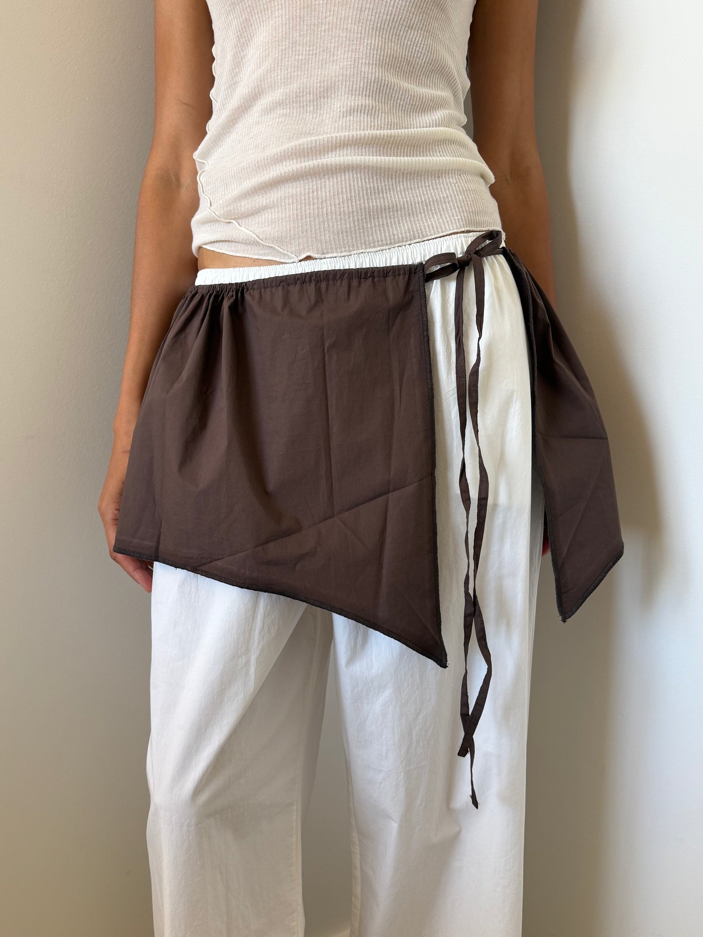 A person wearing a white ribbed tank top and loose organic cotton pants stands against a light wall, styling the Chocolate Napkin Belt from Deiji Studios as an asymmetrical wrap skirt tied around their waist.