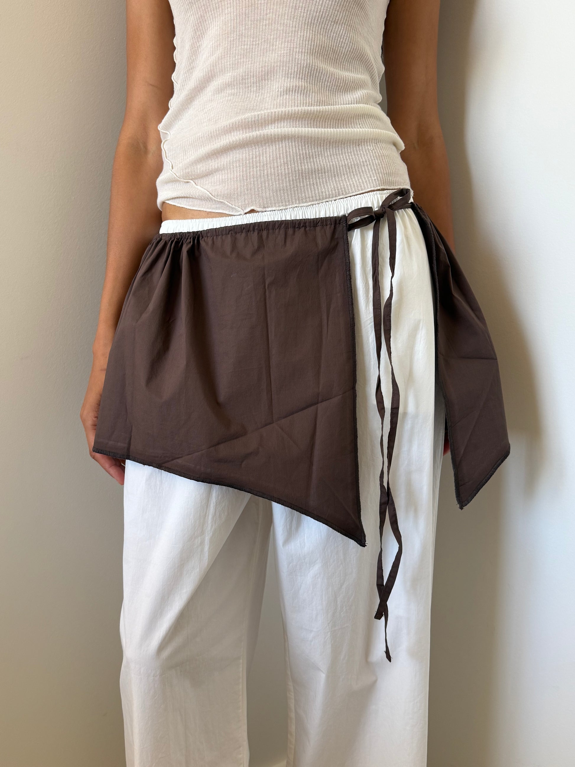 A person wearing a white ribbed tank top and loose organic cotton pants stands against a light wall, styling the Chocolate Napkin Belt from Deiji Studios as an asymmetrical wrap skirt tied around their waist.