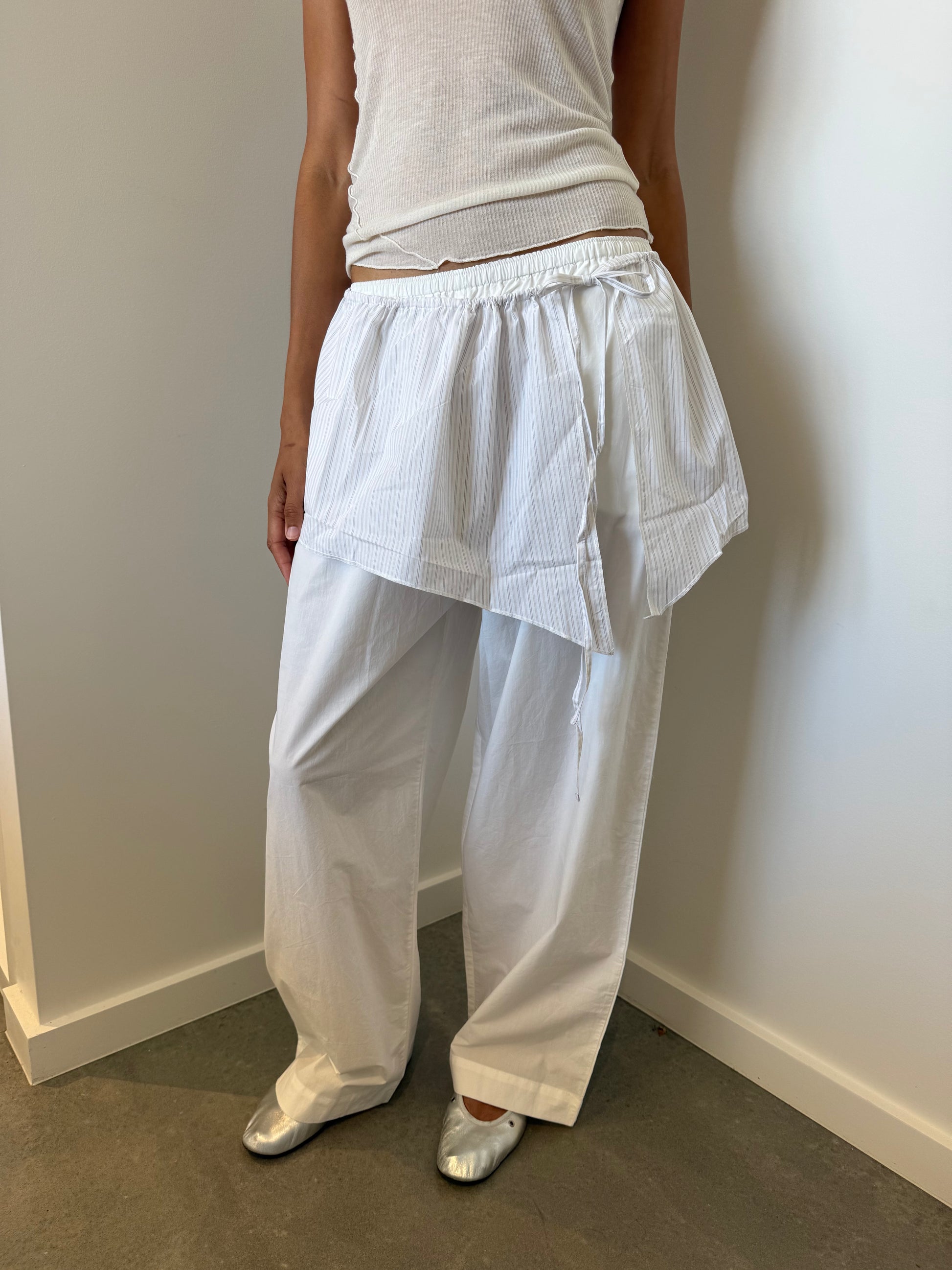 A person wearing a sleeveless white ribbed top, loose organic cotton trousers, a Napkin Belt - Pacific Stripe, and silver flat shoes stands against a white wall on a gray floor.