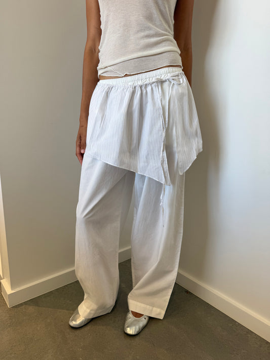 A person stands by a white wall wearing a white sleeveless top, loose organic cotton pants, a white asymmetrical wrap skirt with the Napkin Belt - Pacific Stripe, and silver shoes. Their face is not visible.