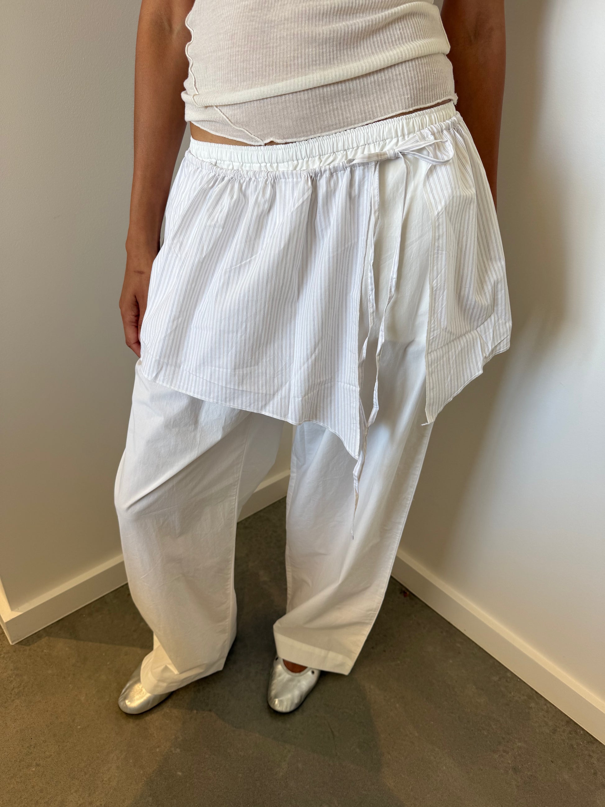 A person stands indoors near a white wall, wearing loose white organic cotton pants, an asymmetrical skirt overlay with a drawstring waist, a Deiji Studios sleeveless top, silver shoes, and the Napkin Belt - Pacific Stripe.
