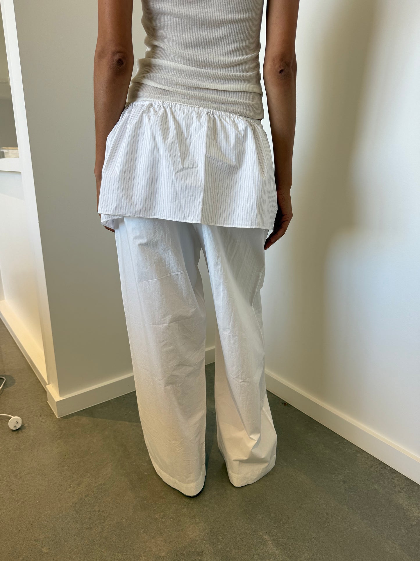A person stands indoors facing away, wearing Deiji Studios' organic cotton white sleeveless top, loose white pants, and a white skirt overlay with the Napkin Belt - Pacific Stripe. The serene setting features white walls and a gray floor.
