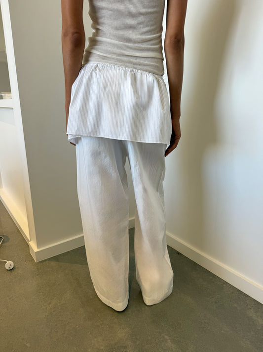 A person stands indoors facing away, wearing a white sleeveless ribbed top, loose organic cotton pants, and the Napkin Belt - Pacific Stripe. The white wall and grey floor evoke Deiji Studios' signature minimalist aesthetic.