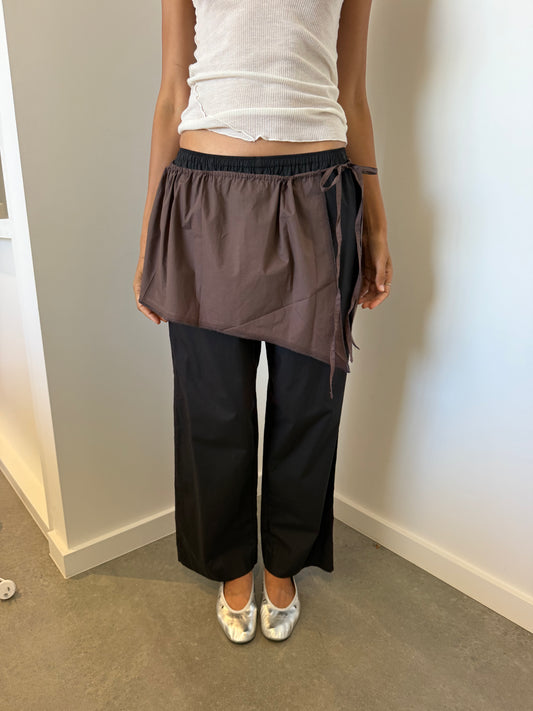A person indoors stands against a light wall, wearing a white organic cotton ribbed tank, black wide-leg pants, silver pointed-toe shoes, and the Napkin Belt - Chocolate as a brown wrap skirt.