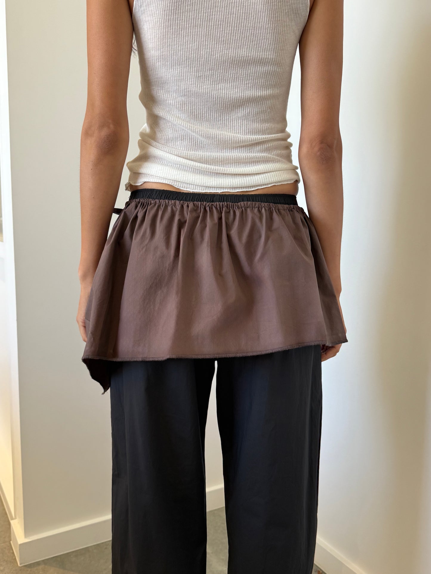 A person stands indoors, seen from the back, wearing a white sleeveless top by Deiji Studios, loose black pants, and the Napkin Belt - Chocolate tied around the waist.