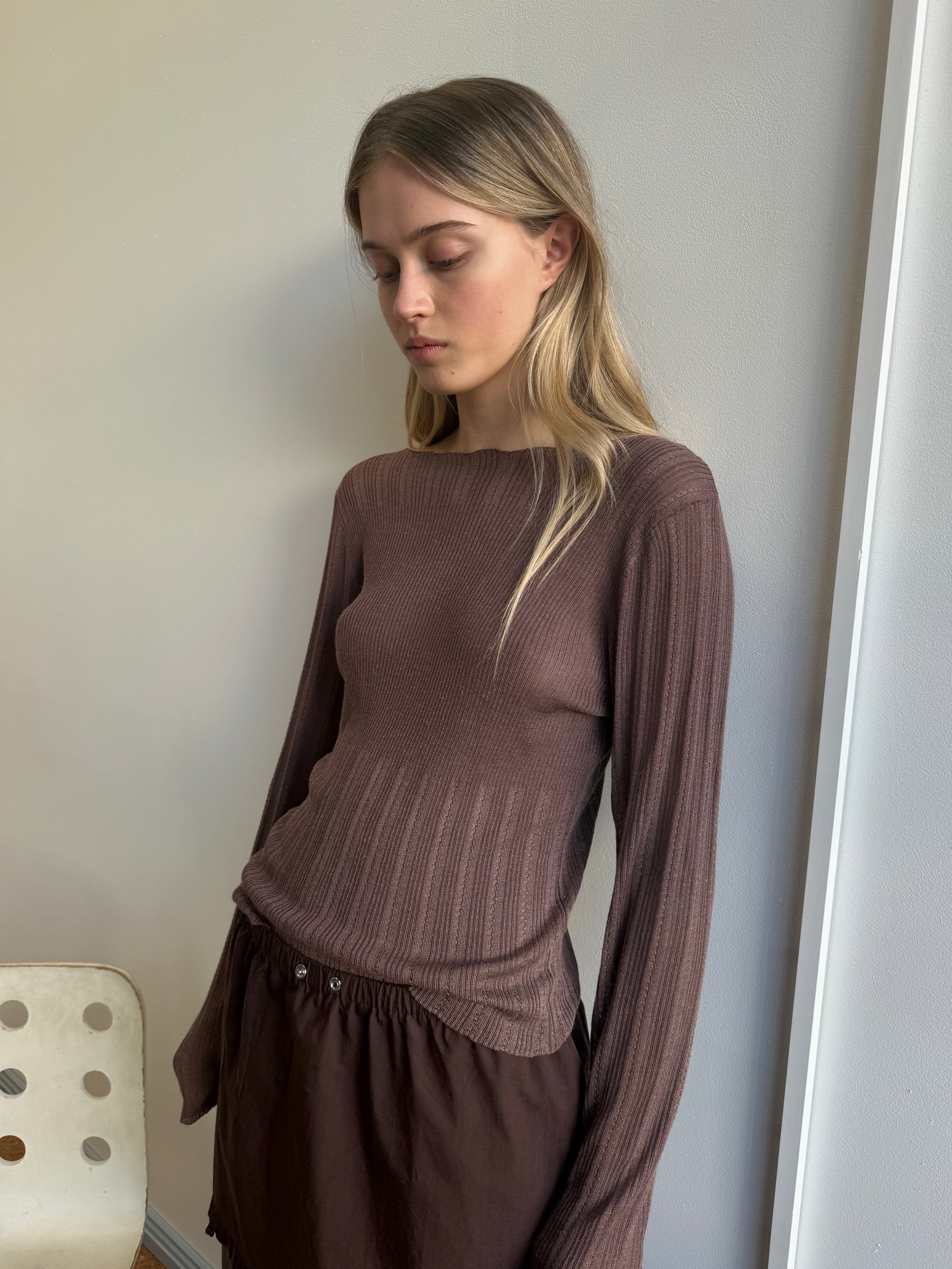 A young woman with long blonde hair stands indoors against a light wall, wearing the Open Work Longsleeve - Hazel and a dark brown skirt. She looks down with a neutral expression.