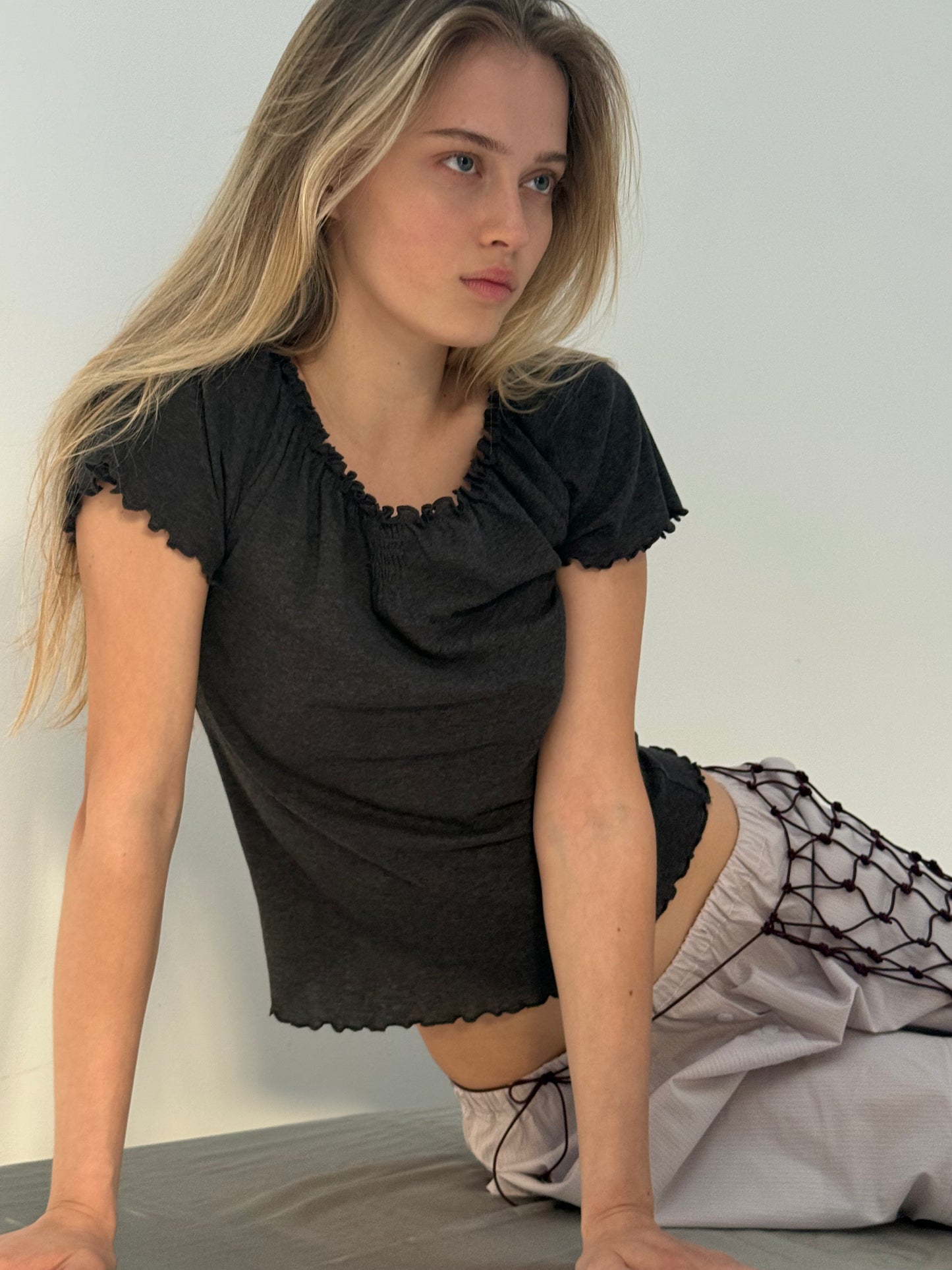 A young woman with long blonde hair sits on a surface wearing the Flounce Tee - Grey by Deiji Studios and light pants with a netted accessory, gazing thoughtfully to the side against a plain, light background.