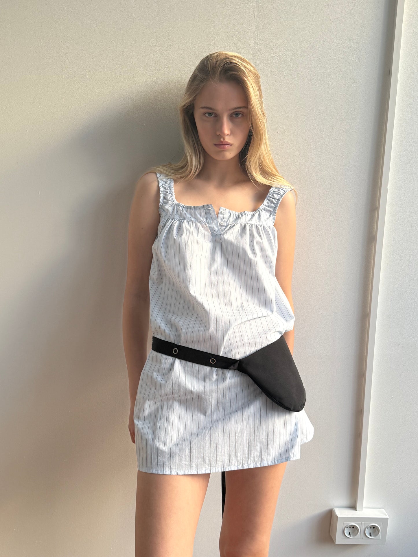 A young woman with long blonde hair stands against a light wall, wearing the sleeveless Yoke Mini Dress - Mineral Stripe by Deiji Studios and a black belt bag. She looks directly at the camera with a serious expression.