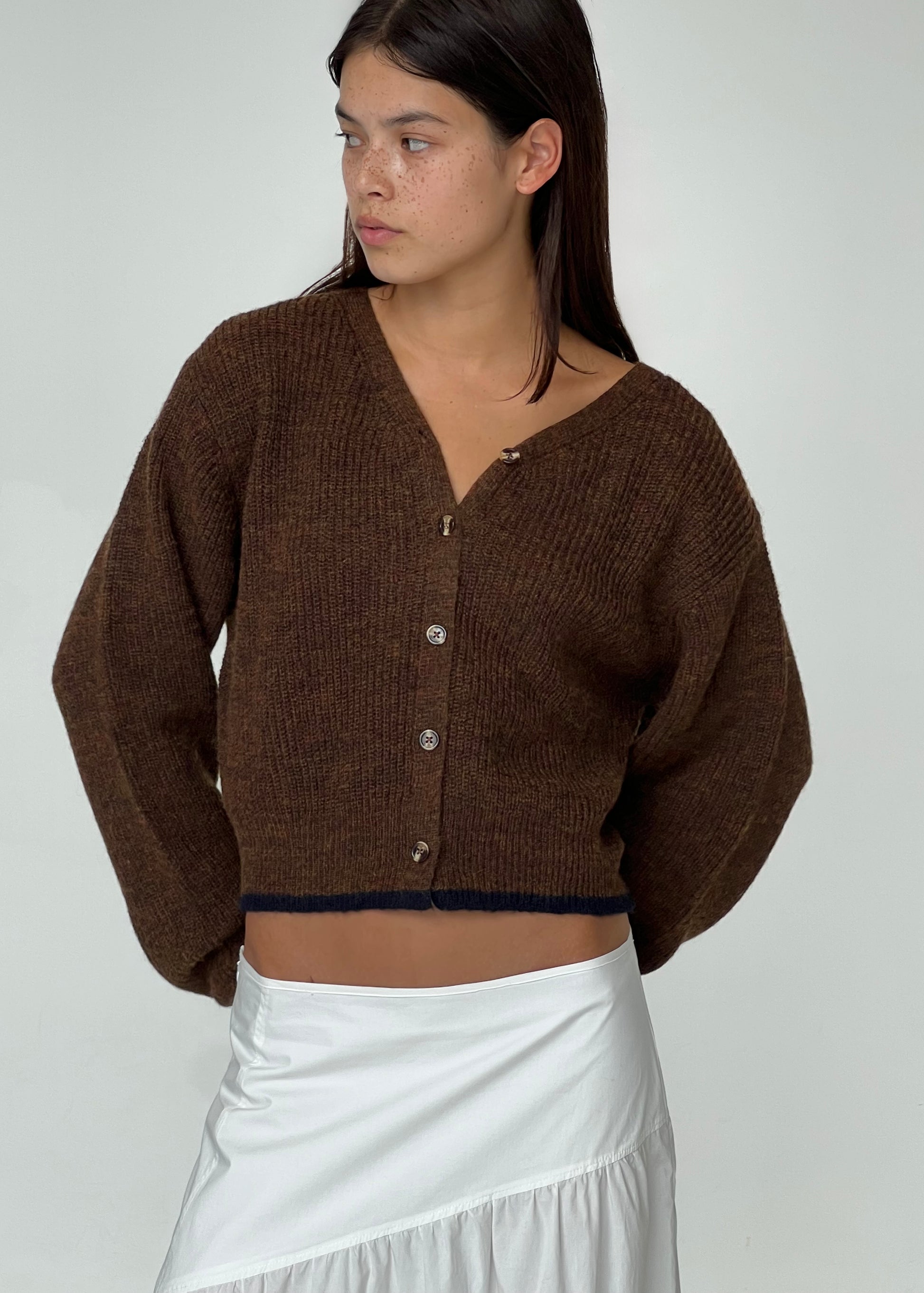 A woman with long dark hair wears the Deiji Studios Cardi - Tawny, a brown sustainable textured knit cardigan, paired with a white skirt. She stands against a plain light background, looking to her left.