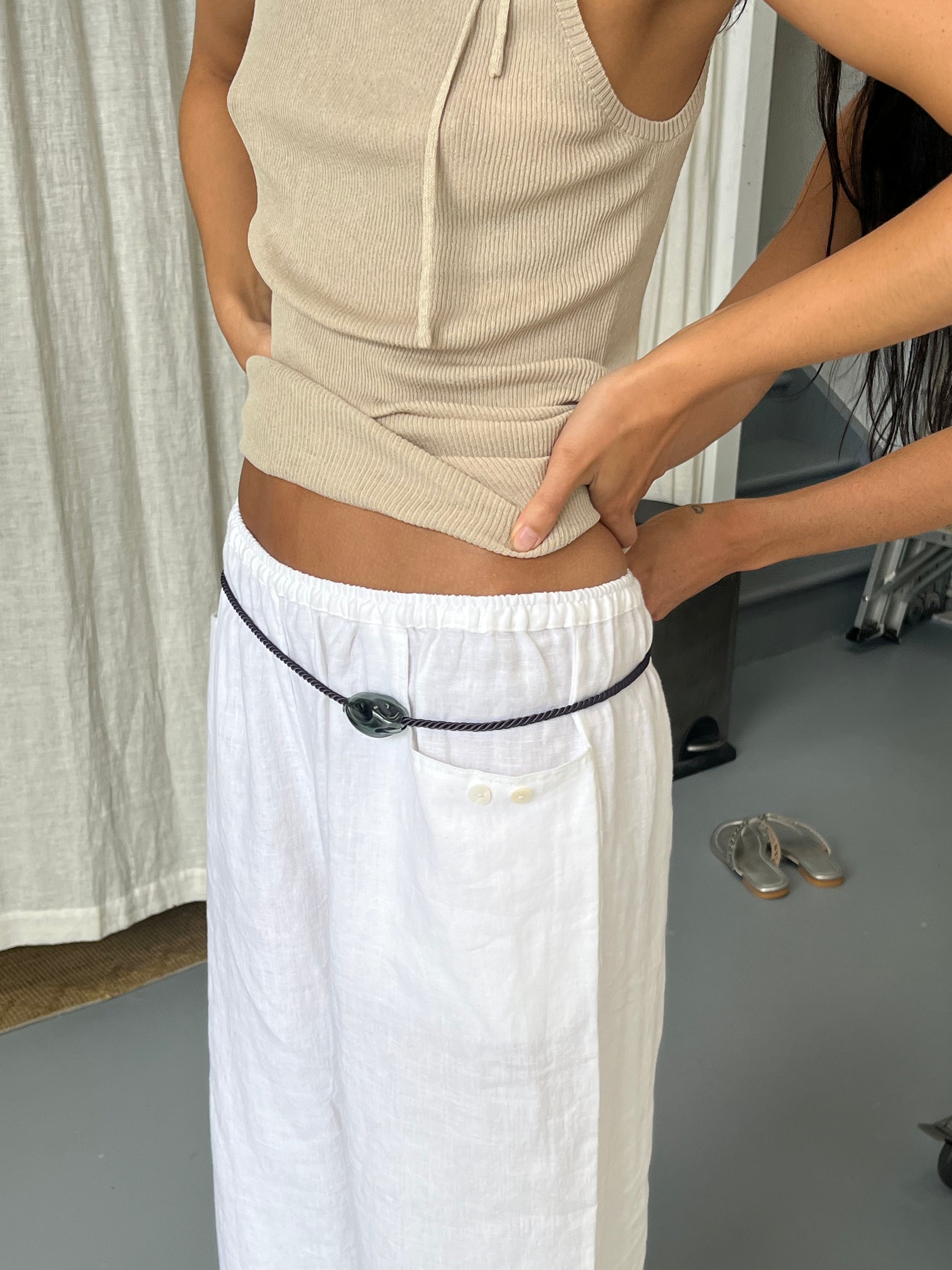 A person wears the Linen Square Pocket Pant - White by Deiji Studios with a beige sleeveless top, cinched by a thin belt with a large stone buckle. Another person adjusts the top while shoes and curtains appear in the background.