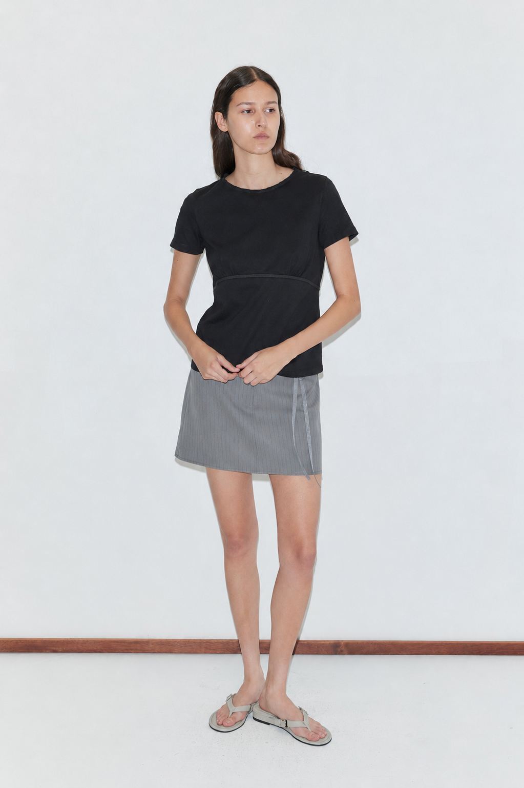 A woman with long dark hair stands against a white background, wearing the Tape Tee in black, a gray pinstriped mini skirt, and flip-flops. Her hands are near her waist.