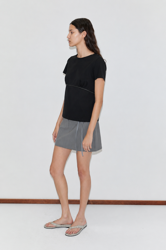 A woman with long brown hair stands sideways against a plain white wall, wearing the Tape Tee - Black, a gray pleated skirt, and beige flip-flops.