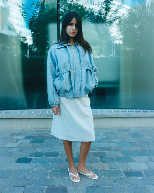A woman stands outdoors on a stone walkway, wearing a light blue oversized denim jacket, the Deiji Studios Simple Skirt in Mint, and white flip-flops. Large glass windows with greenery reflections are in the background.