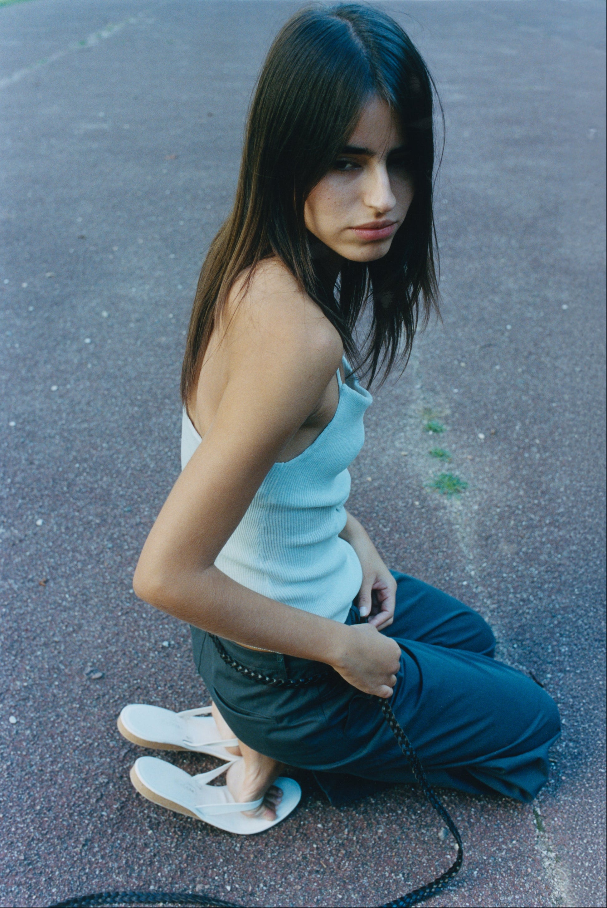 A young woman with long dark hair sits on the ground outdoors, wearing the Deiji Studios Fine Knot Tank in Ice Blue and dark pants with white sandals, her pensive gaze turned slightly away from the camera.