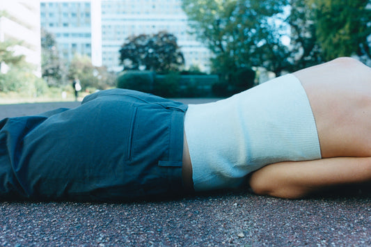 A person wearing dark pants, possibly the Zip Pant - Charcoal, and a light sleeveless top lies on their side outdoors on pavement, with blurred buildings and trees visible in the background.