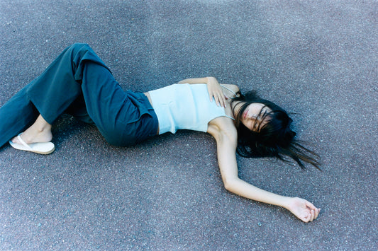 A woman lies on her back on a paved surface, wearing the Fine Knot Tank - Ice Blue by Deiji Studios, paired with dark pants and white shoes. One arm rests on her chest while the other is extended, her hair partially covering her face.
