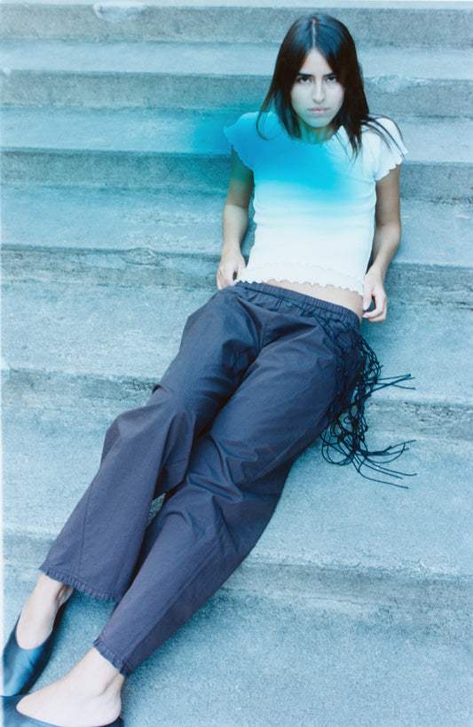 A woman with long dark hair sits on outdoor stone steps, wearing a short-sleeved white top and Deiji Studios' Flounce Pant - Umber, featuring organic cotton and fringe details. She gazes at the camera with a neutral expression.
