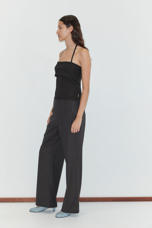 A woman with dark hair stands in profile against a white background, wearing the Half Top - Black, a ribbed knit halter top, paired with dark pinstriped wide-leg trousers and light blue heeled shoes.