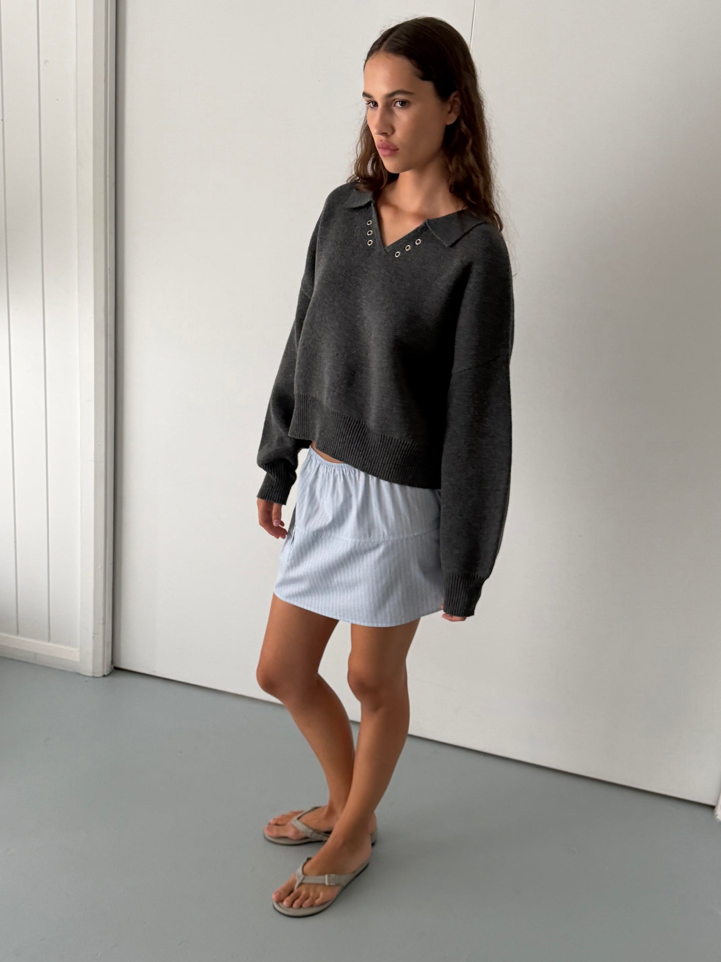 A woman with long brown hair stands indoors against plain white walls, wearing the Eyelet Knit - Dark Pebble sweater from Deiji Studios, a light blue skirt, and beige sandals. She gazes slightly to the side.
