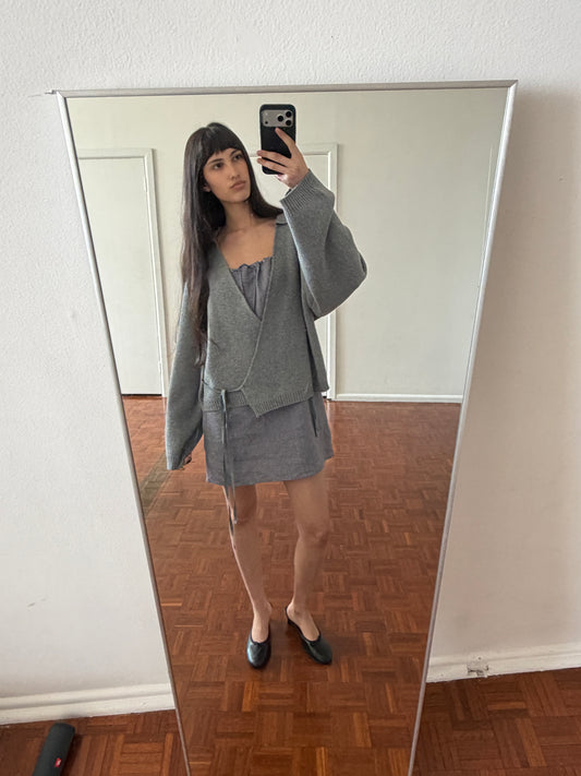 A woman with dark hair and a fringe takes a mirror photo wearing the Wrap Cardigan - Grey by Deiji Studios, a grey knit wrap cardigan with a waist tie, over a grey top and mini skirt with black flats.