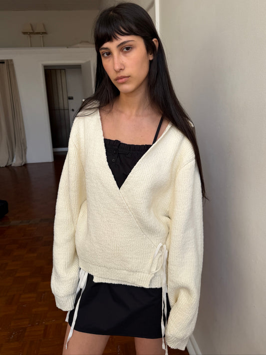 A woman with dark hair and a fringe stands in a room wearing the Wrap Cardigan - Snow by Deiji Studios, a cream knit wrap cardigan with a collar and waist tie, over a black top and mini skirt.