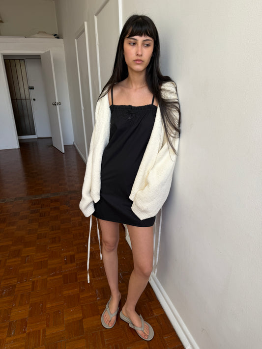 A woman with dark hair and a fringe leans against a wall wearing the Wrap Cardigan - Snow by Deiji Studios, an oversized cream knit wrap cardigan, over a black slip dress with flip-flops.