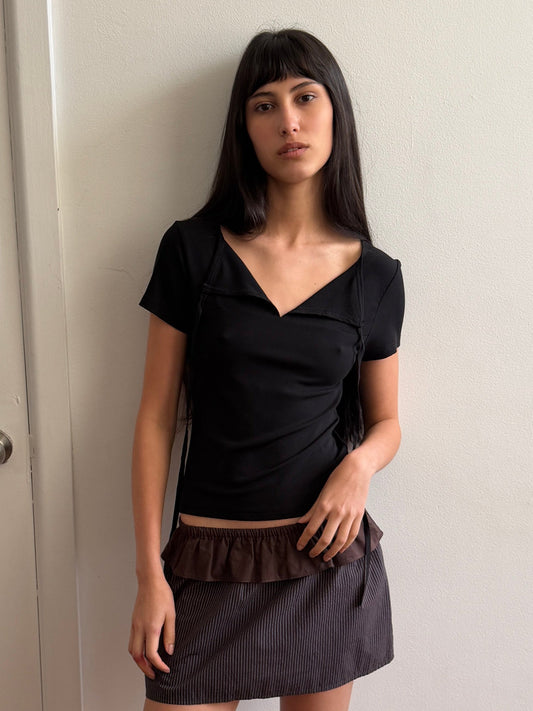 A woman with dark hair and a fringe leans against a wall wearing the Tie Back Tee - Black by Deiji Studios, a fitted black tee with a sweetheart neckline and back ties, with a brown flounce mini skirt and flip-flops.