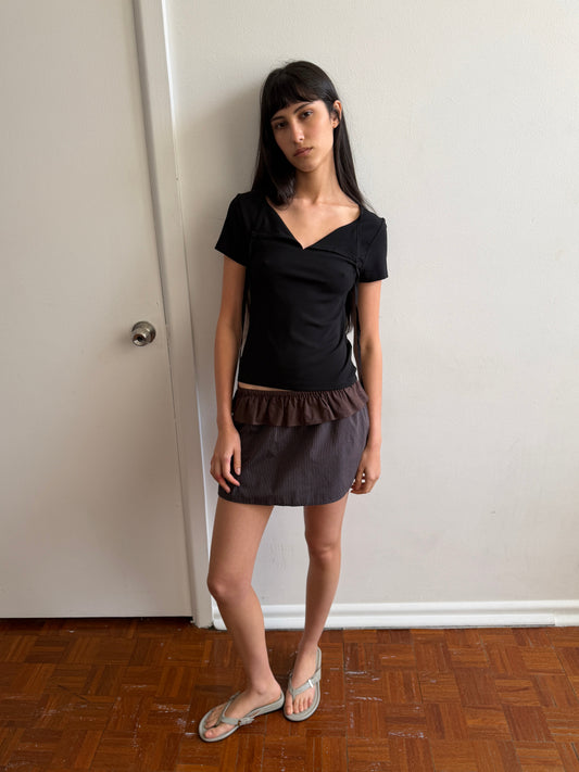 A woman with dark hair and a fringe stands against a door wearing the Tie Back Tee - Black by Deiji Studios, a fitted black tee with a sweetheart neckline, with a brown flounce mini skirt and flip-flops.