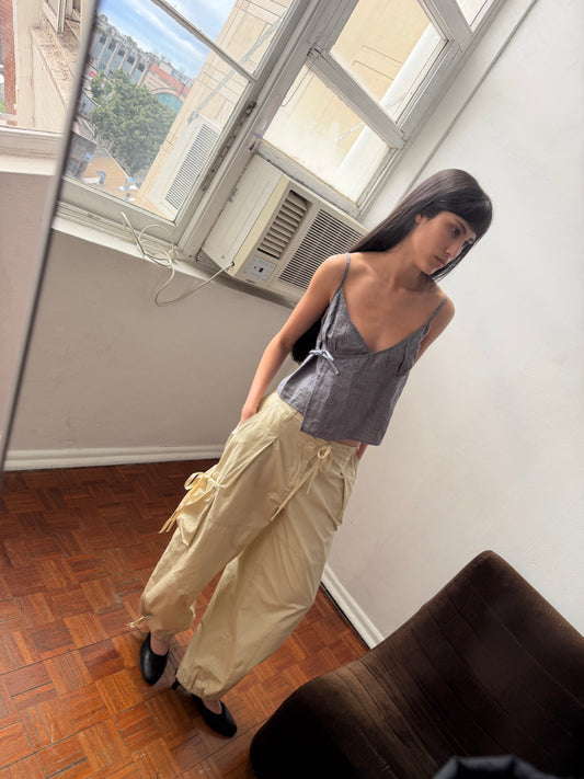 A woman with dark hair and a fringe stands in a room wearing the Wrapping Top - Indigo Check by Deiji Studios, an indigo micro-check linen cami with a wrap front and side tie, with cream drawcord trousers.