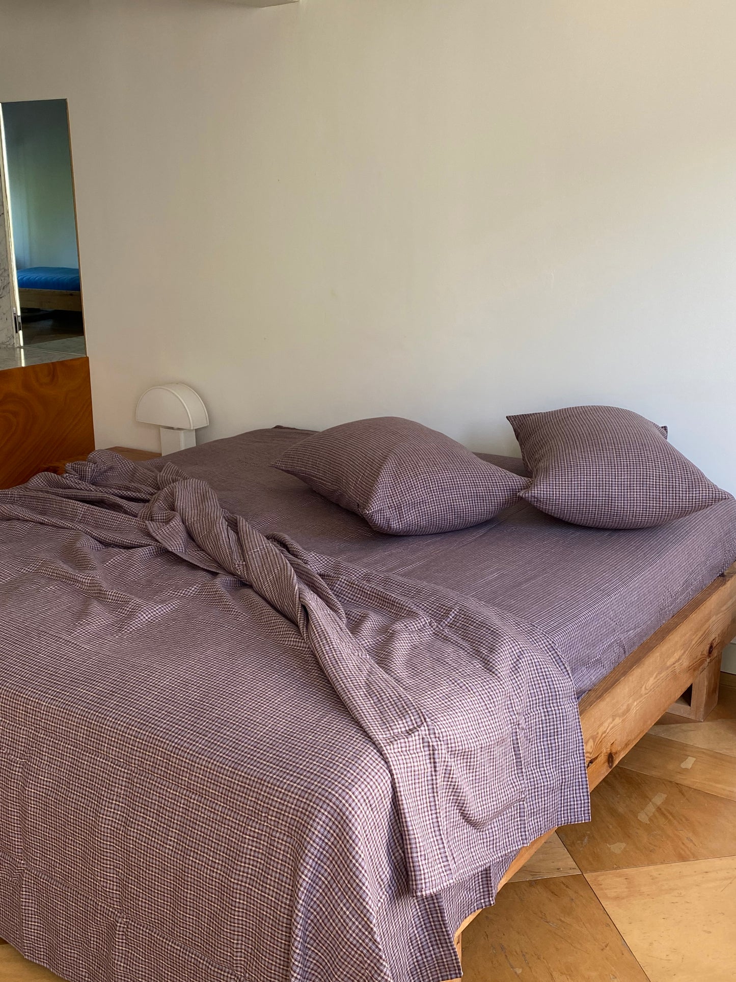 A neatly made wooden bed is dressed in purple checkered sheets and two Pillow slip set - Russet Check. The sheet is slightly rumpled, with a small white lamp on the bedside table against a plain wall.