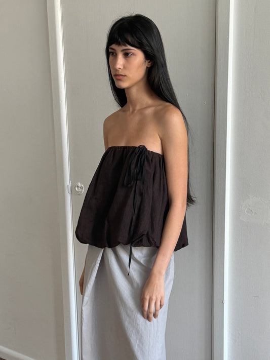 A woman with dark hair and a fringe stands in profile in a room wearing the Bubble Top - Brown Black Stripe by Deiji Studios, a strapless dark brown bubble-hem top with a drawcord tie, over a pale grey skirt.